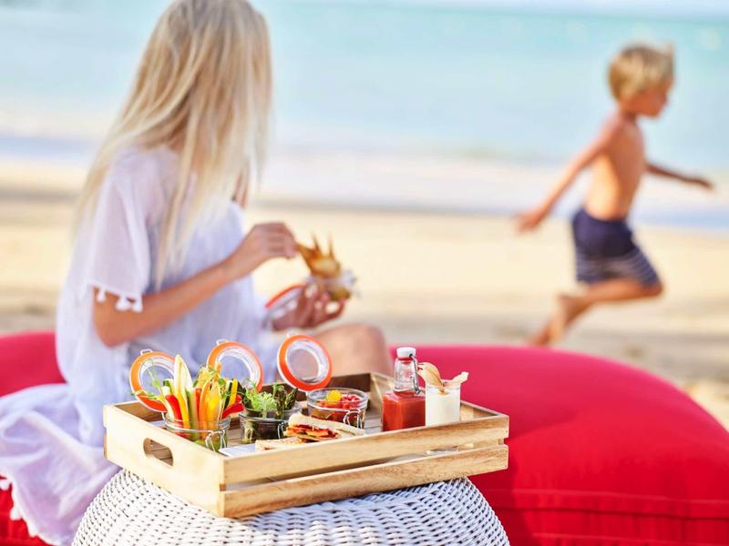 Vrouw houdt dienblad met ontbijtopties op het strand, kind rent op de achtergrond.