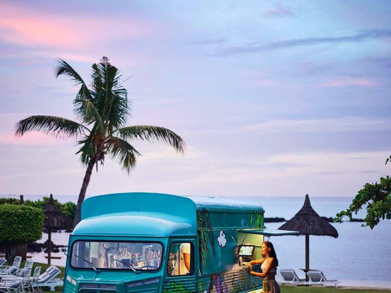 Vrouw staat naast een kleurrijke bus op gras met palmbomen en zee op de achtergrond.