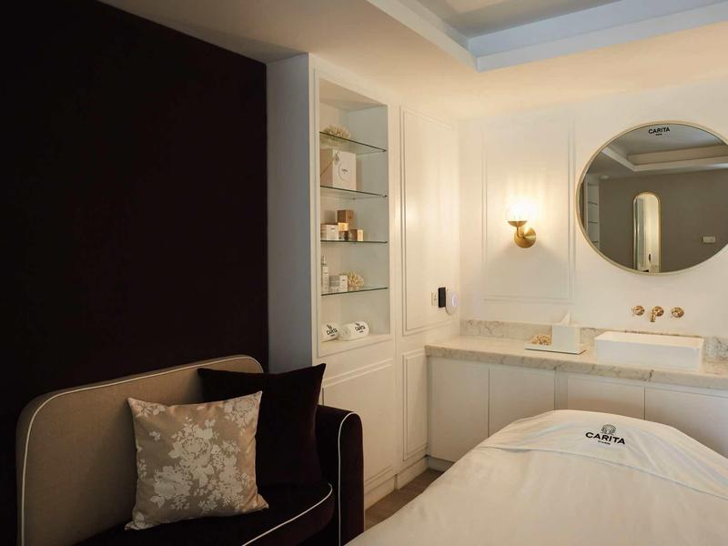 Comfortabele behandelruimte met ligstoel, bank en moderne wastafel in neutrale kleuren.