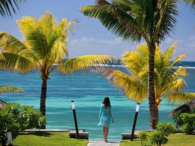 Žena v modrých šatech se dívá na tropické moře obklopené palmami a zeleným trávníkem
