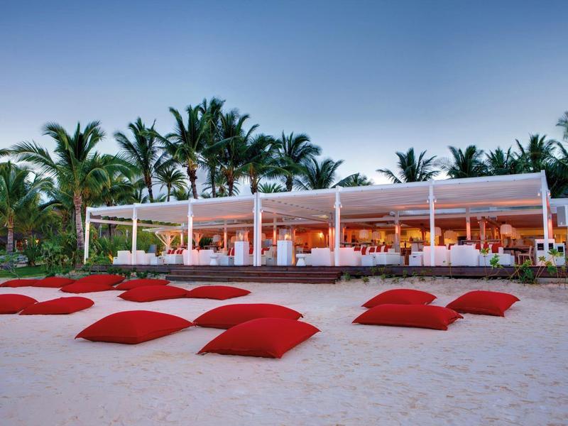 Plážová restaurace s bílými stoly, židlemi a červenými polštáři na písku před palmami.