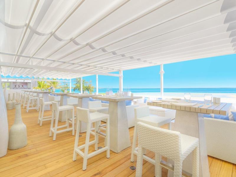 Světle zařízený plážový bar s bílým nábytkem a výhledem na moře pod modrou oblohou.