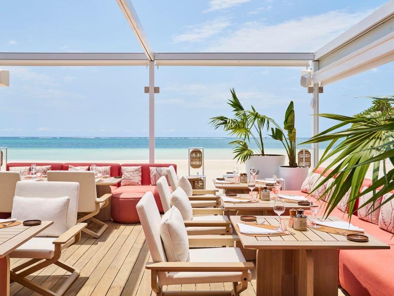 Světelná plážová restaurace s bílými židlemi, dřevěnými stoly a výhledem na moře pod jasnou oblohou.