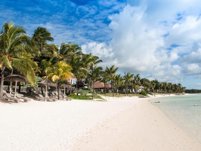 Bílá písečná pláž s palmami a slunečníky pod modrou oblohou u moře.