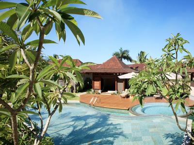 Een tropisch resort met een zwembad, omringd door palmbomen en bungalows onder een heldere blauwe lucht.