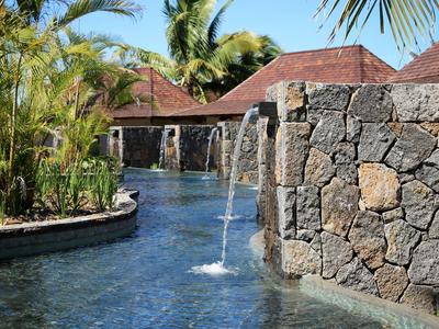 Een zwembad met watervallen, omgeven door stenen muren en tropische planten onder een blauwe lucht.