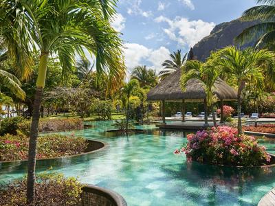 Tropischer Poolbereich mit Palmen und einem überdachten Sitzbereich unter blauem Himmel.