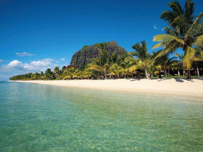 Heldere oceaan met wit zandstrand, palmbomen en een berg op de achtergrond onder blauwe hemel.