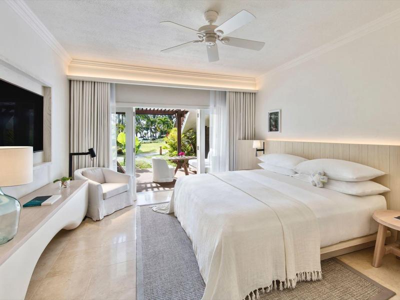 Helder hotelkamer met tweepersoonsbed, fauteuils en uitzicht op een tuin met zwembad.