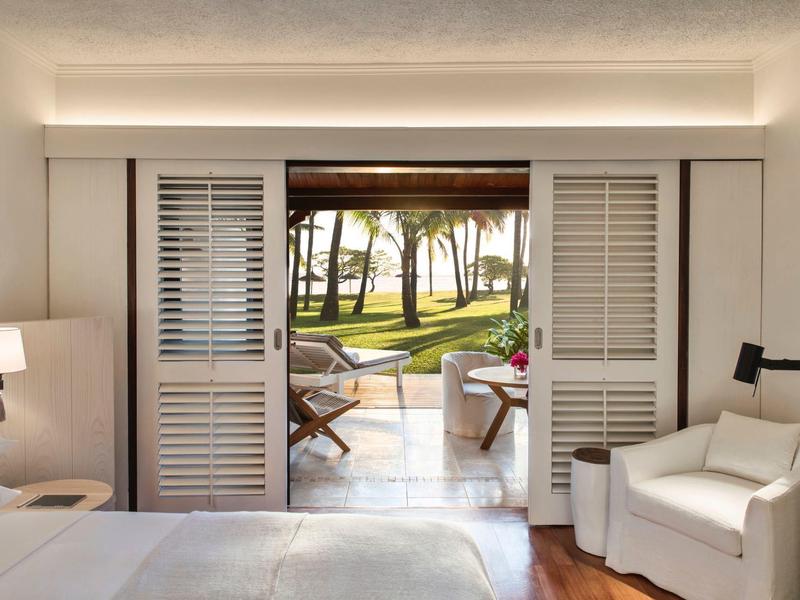 Uitzicht vanuit een hotelkamer met wit meubilair op een terras en palmbomen in de tuin.