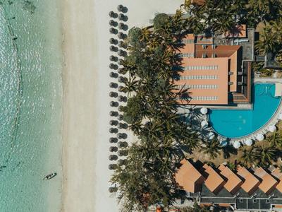 Luftaufnahme eines Resorts mit Pool, Palmen, Liegestühlen und weißem Sandstrand am klaren Wasser.