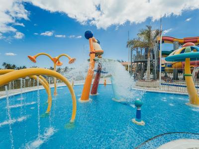 Wasserpark mit Wasserrutschen, Wasserspielen und einem Planschbecken unter blauem Himmel.