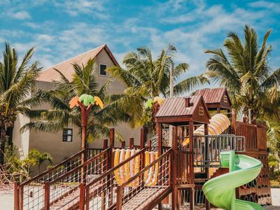 Spielplatz mit Rutschen und Wippen vor einem Haus und Palmen unter blau bewölktem Himmel.