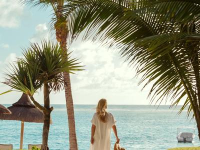 Eine Frau in einem weißen Kleid geht mit einem Kind am Strand unter Palmen spazieren.