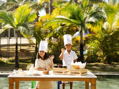 Zwei Köche mit Kochmützen bereiten an einem Tisch im Freien Essen vor, umgeben von Palmen.