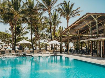 Schwimmbad mit klarem Wasser, umgeben von Palmen und Sonnenschirmen neben einer überdachten Terrasse.