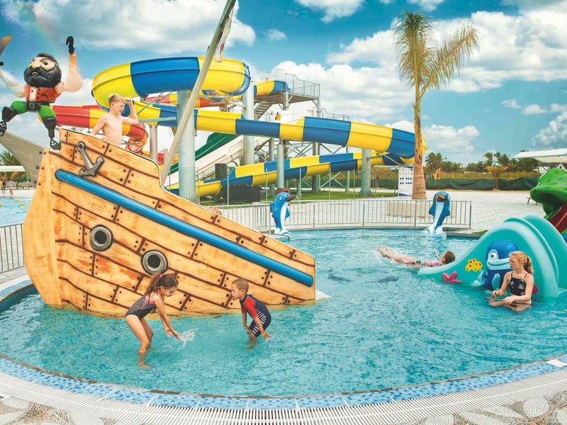 Kinderen spelen in een zwembad met een piratenschip en glijbanen onder een gedeeltelijk bewolkte hemel.