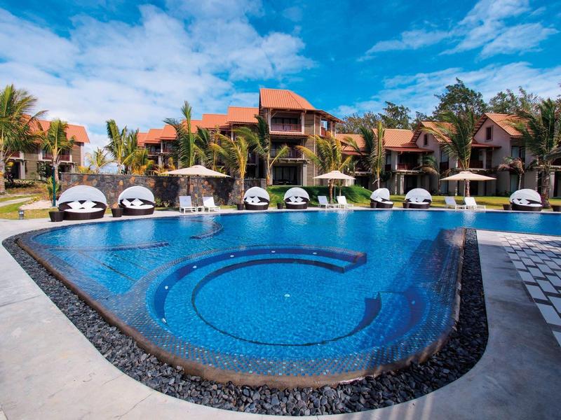 Resortzwembad met parasols en ligbedden, omringd door tropische villa's onder een blauwe lucht.