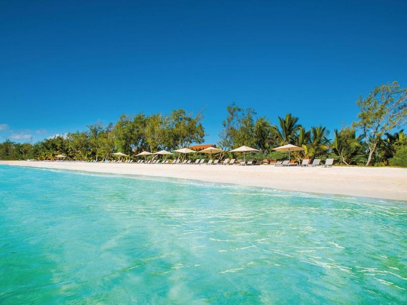 Helder turquoise water grenst aan een zandstrand met parasols en groene bomen onder een blauwe lucht.