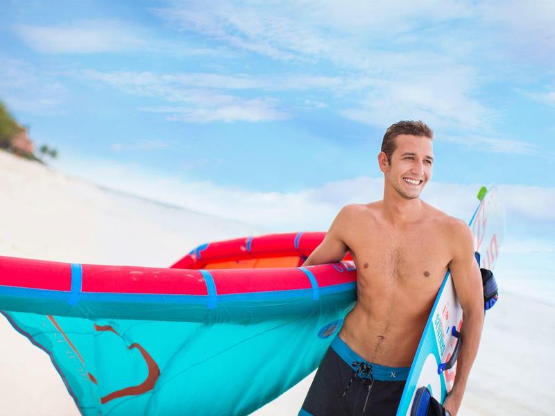 Glimlachende man zonder shirt met een kleurrijk kitesurfzeil op een zandstrand onder een blauwe lucht.
