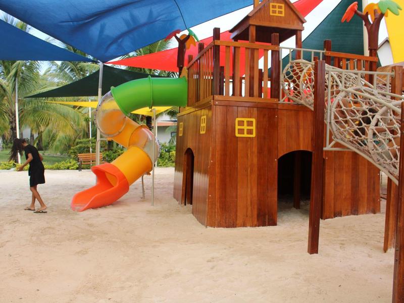 Buitenspeeltuin met houten kasteel, glijbanen, klimnet en spelende kinderen op zand.