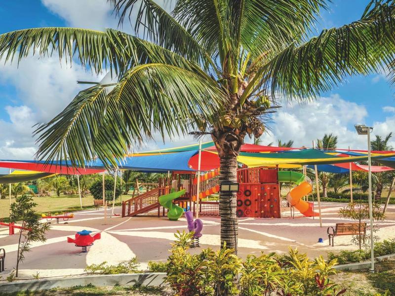 Buitenspeeltuin met kleurrijke schaduwzeilen en een palmboom onder een blauwe hemel.