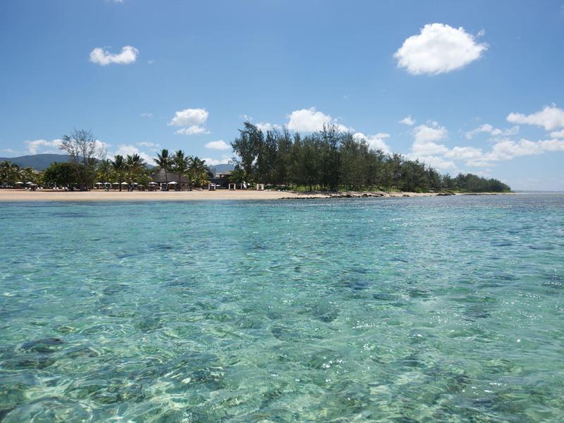 Klares türkisfarbenes Wasser trifft auf einen bewaldeten, sandigen Strand unter blauem Himmel mit Wolken.
