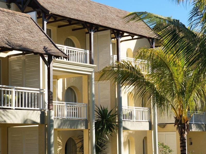 Gelbes zweistöckiges Haus mit weißen Balkonen, umgeben von Palmen und grünem Garten unter blauem Himmel.