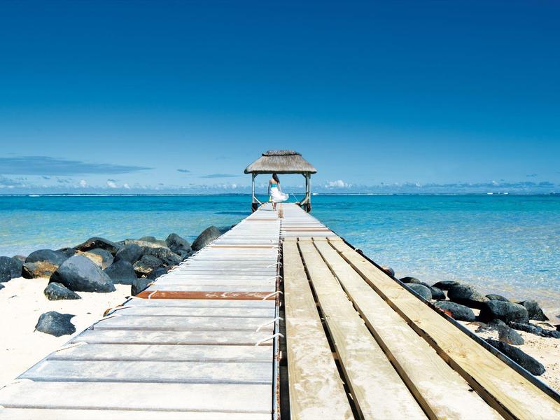 Holzsteg mit Sonnenschirm mit weißen Stühlen endet am klaren blauen Meer unter klarem Himmel.