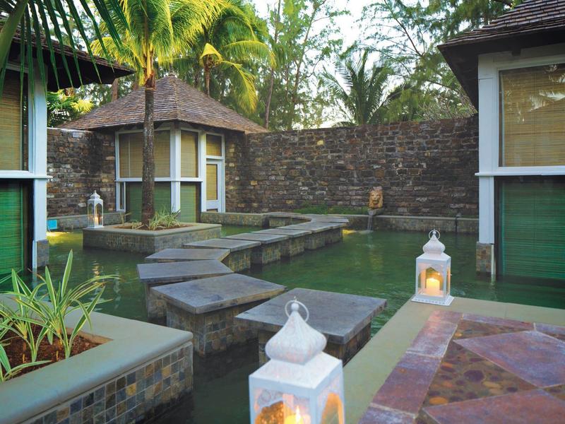 Ein Garten mit Wasserbecken, Steinstufen und kleinen grünen Gebäuden, umgeben von Palmen.
