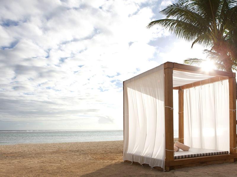 Strand mit Holzbett, weißen Vorhängen, Palmen und bewölktem Himmel bei Sonnenlicht.