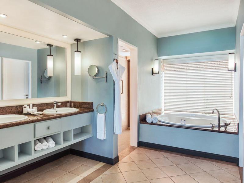 Helles Badezimmer mit blauem Wandton, großer Spiegelwand, Doppelwaschbecken und Badewanne unter Fenster.