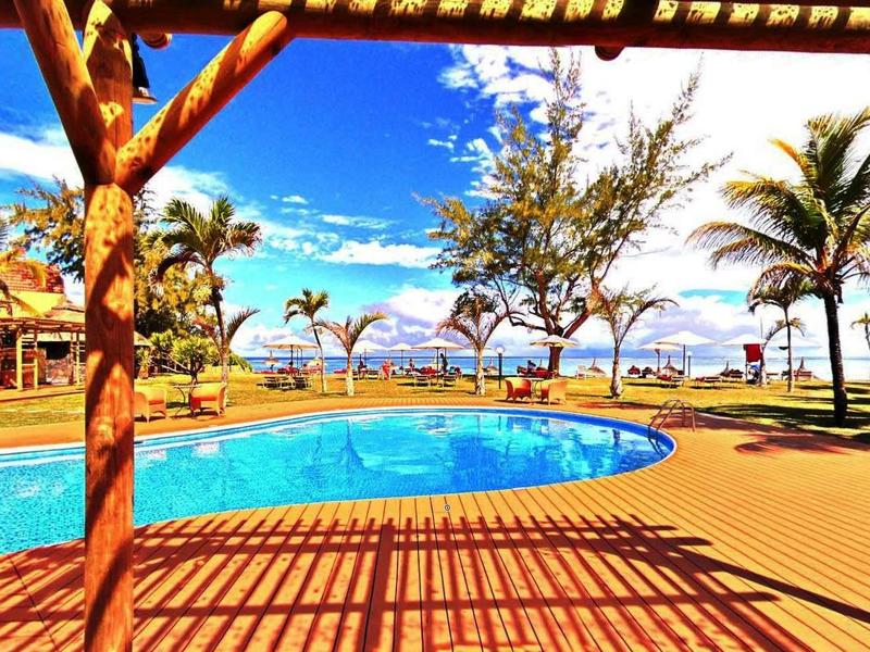 Area piscina di un resort con palme, pergolato in legno e cielo azzurro brillante.