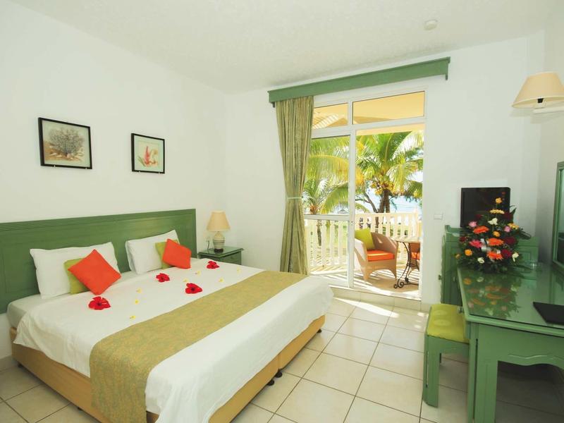 Camera d'hotel luminosa con letto matrimoniale, mobilia verde e balcone con vista sulle palme.