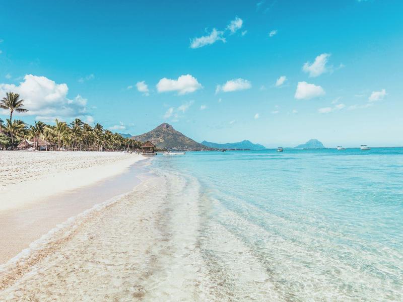 Helder turquoise water spoelt tegen een zandstrand met palmbomen en verre heuvels.