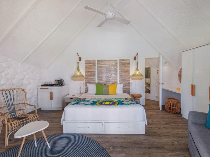 Gezellige slaapkamer met wit bed, kleurrijke kussens, rotan stoel en houten vloer in een lichte kamer.