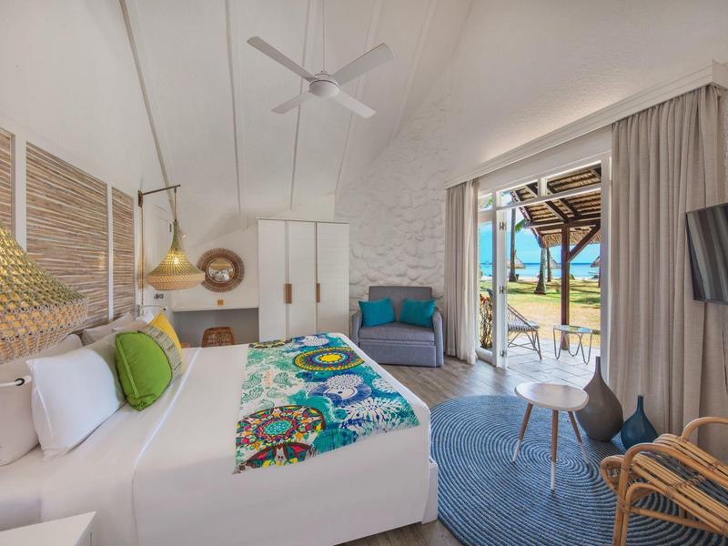 Lichte hotelkamer met wit bed, kleurrijke kussens, blauwe stoel en uitzicht op strand door open deur.