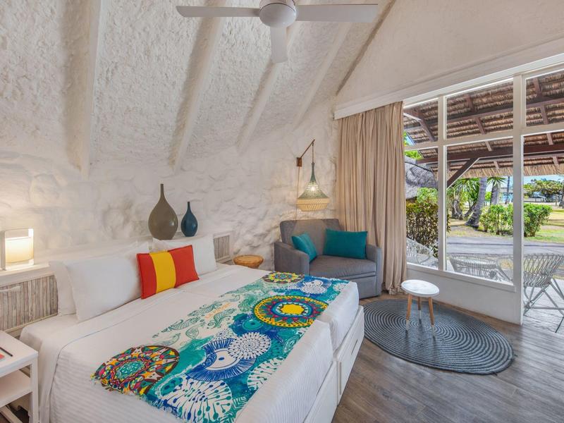 Gezellige kamer met witte stenen muren, bed met kleurrijk deken, zithoek en patio met tuinzicht.