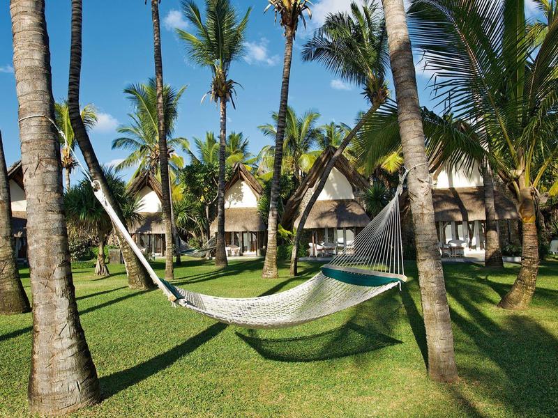 Hangmat tussen palmbomen op groen grasveld bij bungalows onder blauwe lucht.