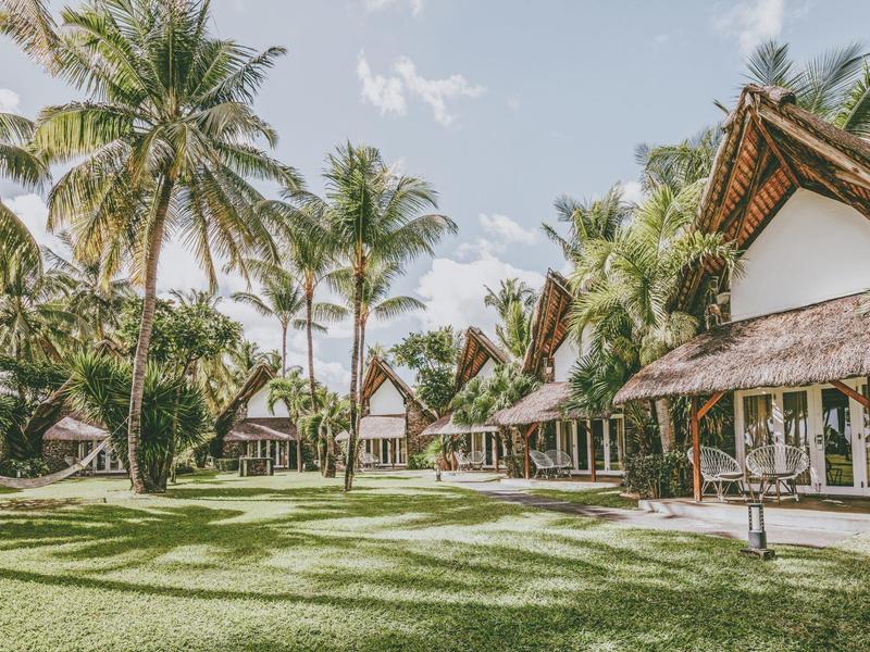 Tropisch resort met houten huisjes, palmbomen en groen gras onder een blauwe lucht.