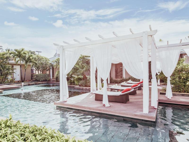 Buiten cabana met witte gordijnen naast een zwembad en groen onder een blauwe lucht.