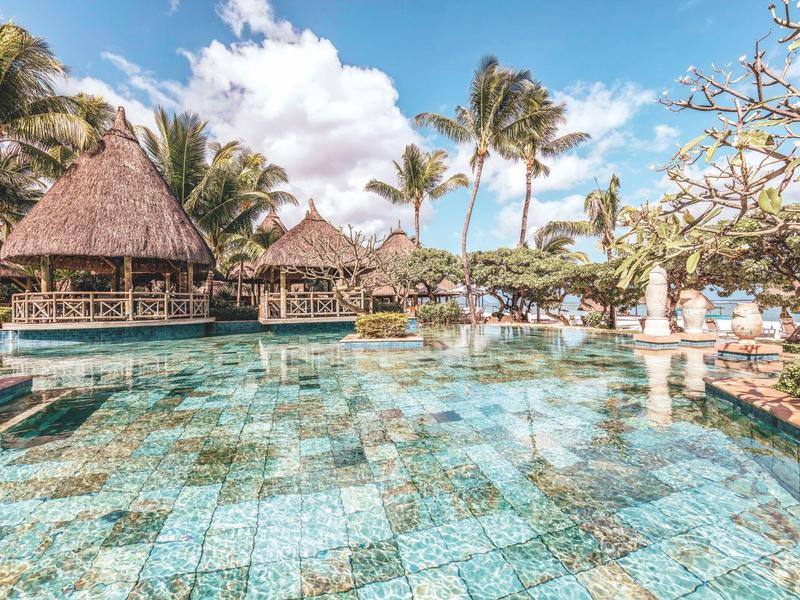 Resortzwembad met turkooizen tegels omgeven door palmbomen en rieten bungalows onder een blauwe lucht.