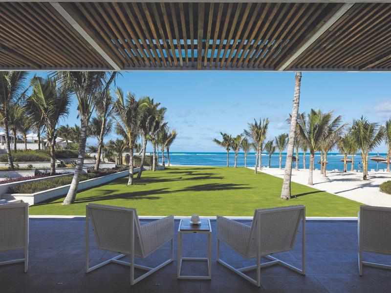 Buiten zitgedeelte met uitzicht op palmbomen, groen gazon en oceaan onder helderblauwe lucht.