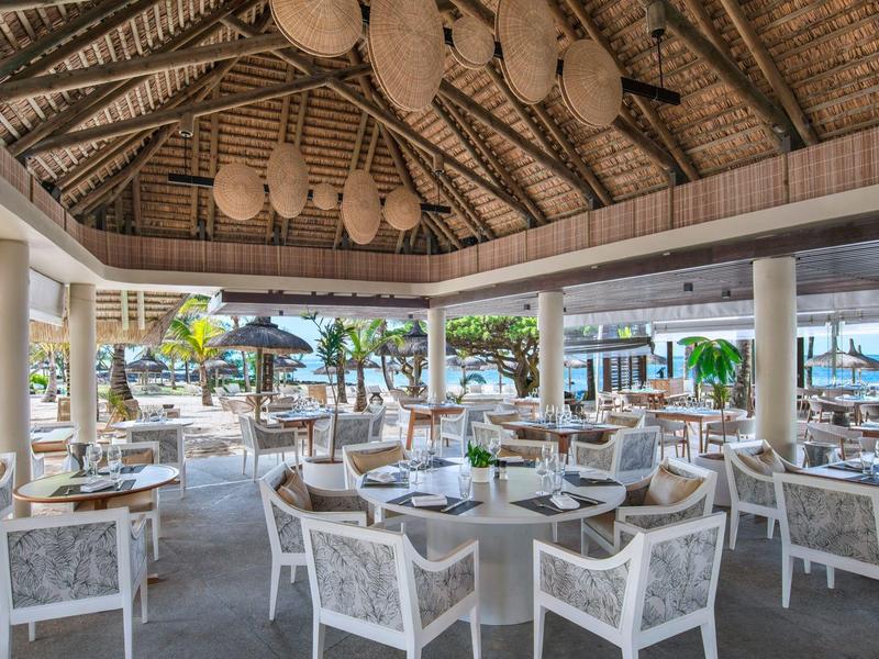 Openluchtrestaurant onder een rieten dak met witte stoelen en houten tafels vlakbij het strand.