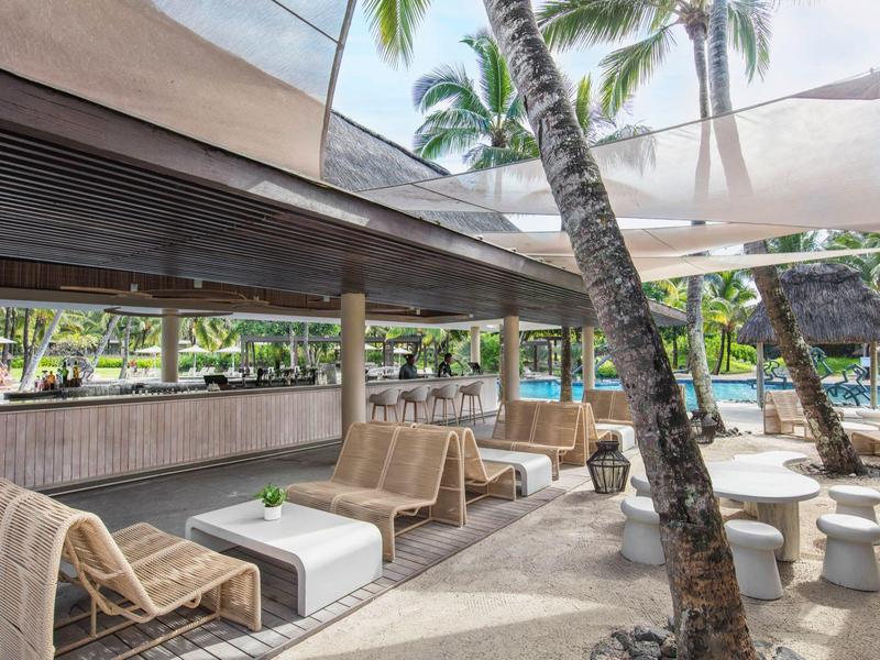 Buitenlounge met banken, schaduwdoeken, palmbomen en zwembad in een tropische omgeving.