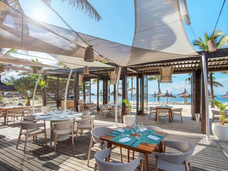 Buitenterras aan het strand met houten vloer, tafels, stoelen en schaduwzeilen bij palmbomen.