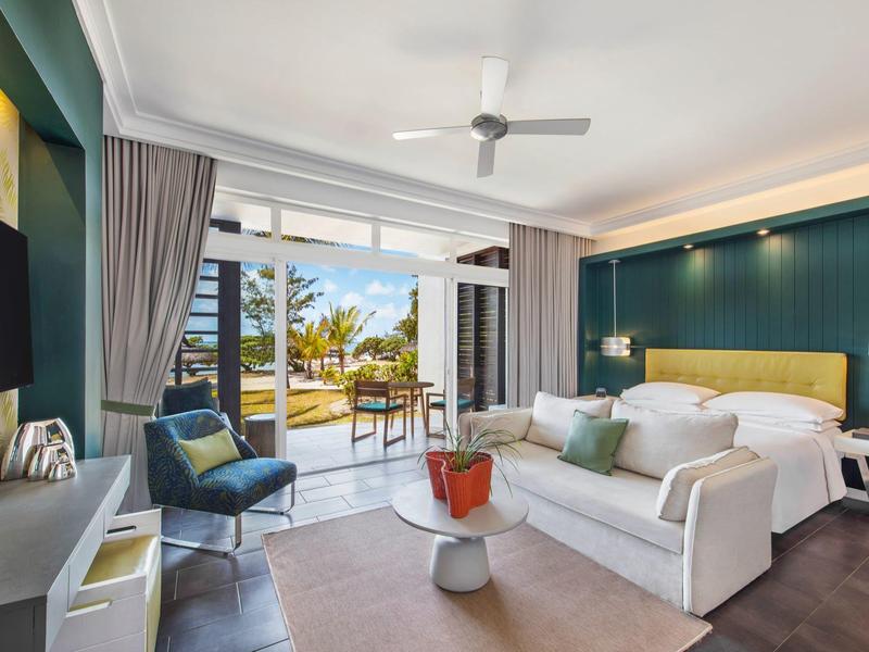 Moderne hotelkamer met bank, fauteuil, tv, schuifdeuren naar zonnig terras met zitgelegenheid.