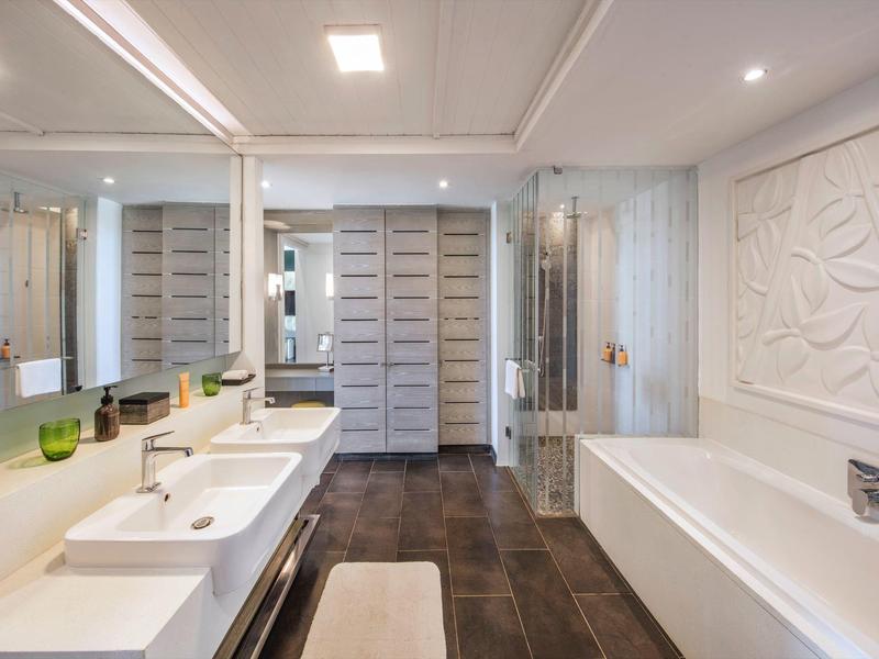 Moderne badkamer met dubbele wastafels, groot bad, gestructureerd wit wandpaneel en donkere vloertegels.