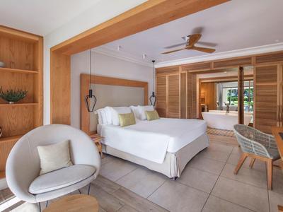 Habitación de hotel luminosa con cama doble, área de estar, ventilador y muebles de madera, abierta al baño.