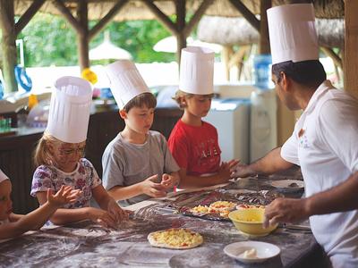 Niños con gorros de chef preparando pizza en una cocina exterior cubierta con un chef.